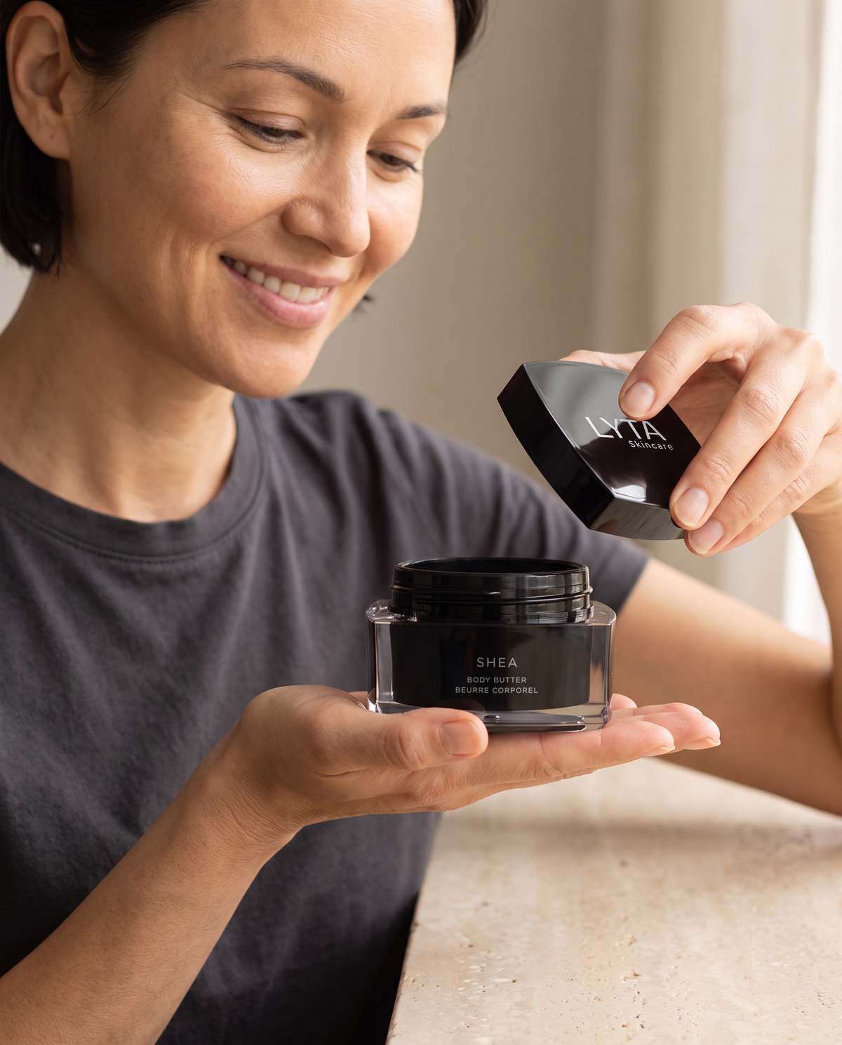 LYTA Shea Body Butter, hands holding the original clear-edged jar and rounded square black lid